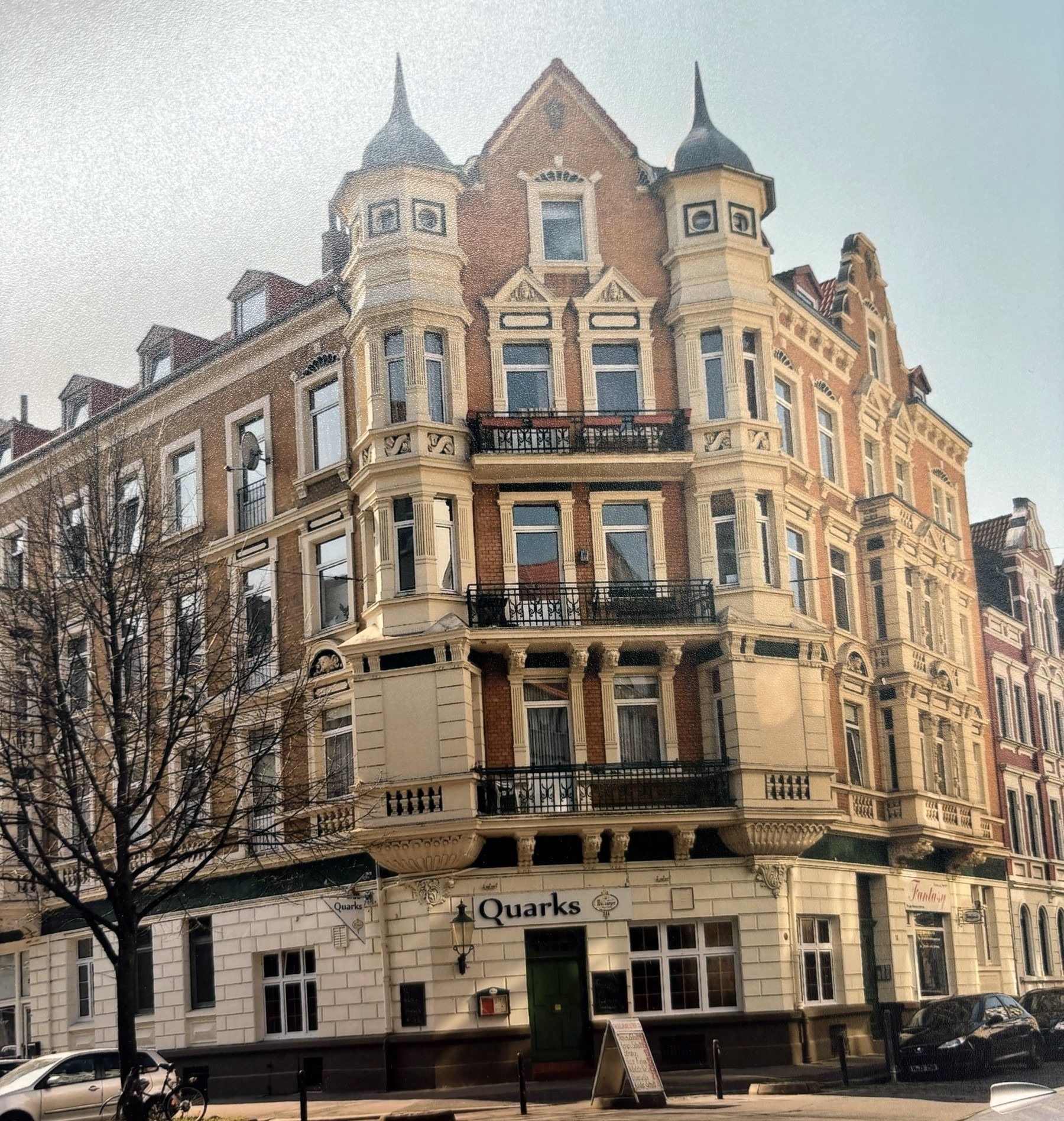 Farbfoto des Gründerzeit-Eckhauses Seydlitzstraße 34: sandfarbene Fassade, zwei spitze Ecktürme, schmiedeeiserne Balkone, im Erdgeschoss das Lokal „Quarks“